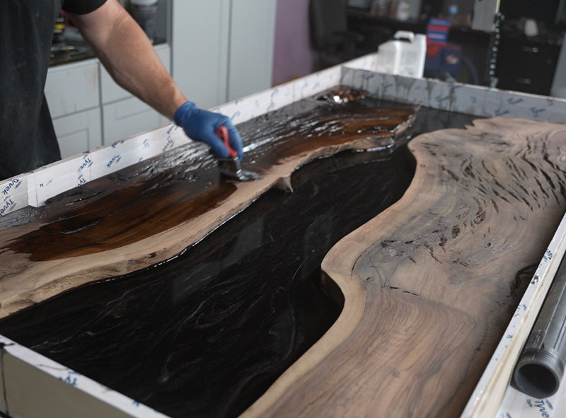 Person working on a large wooden surface, such as a river table, with penetrating epoxy resin, likely in a workshop setting.