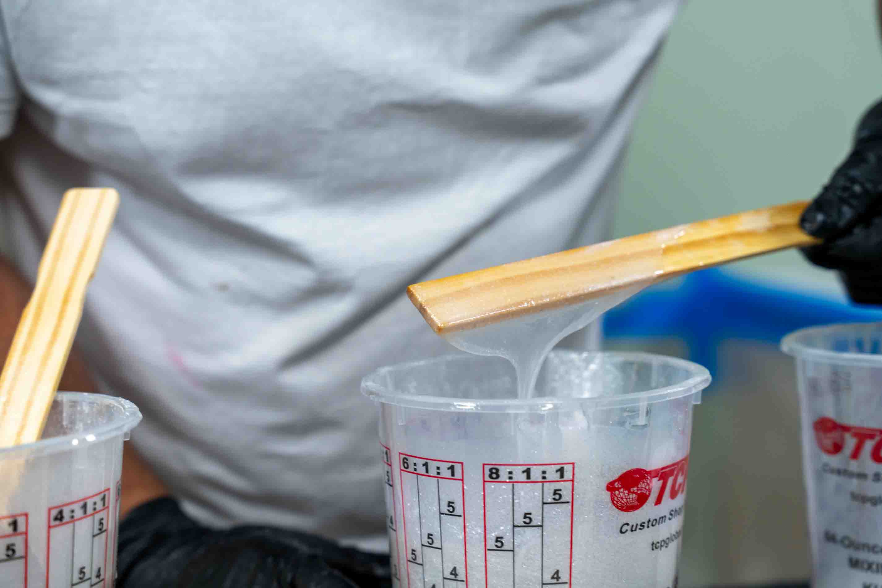 A close-up of a person mixing a translucent white polyaspartic in clear plastic measuring cups using wooden sticks, with one hand wearing a black glove.