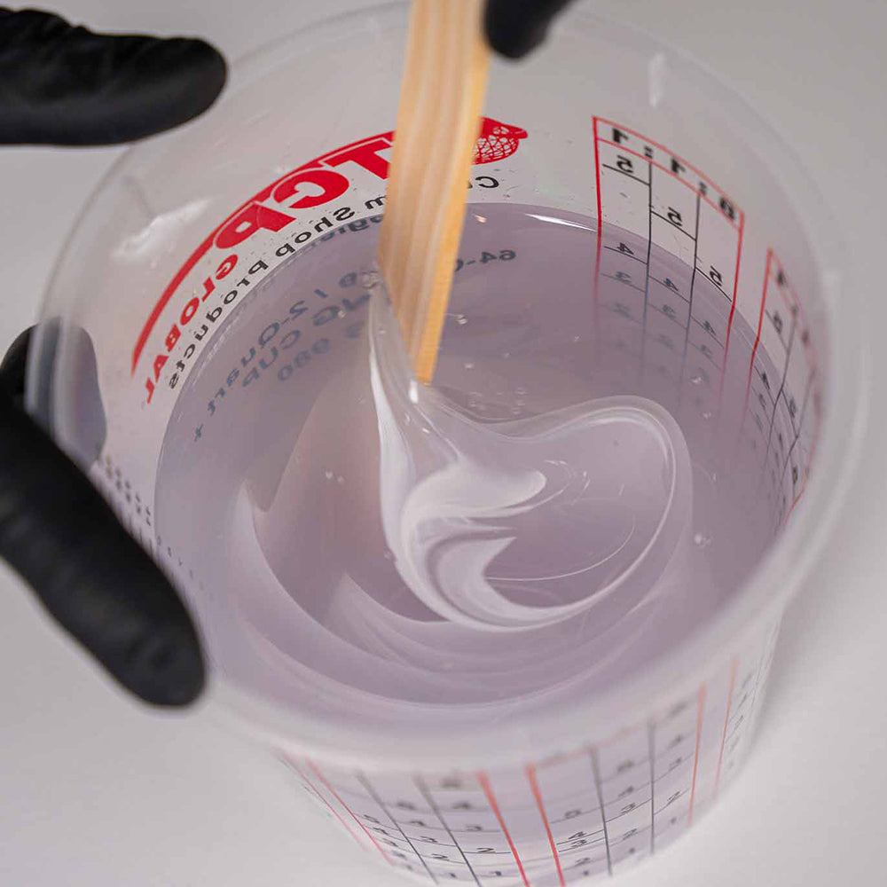 Measuring cup with polyaspartic and white dye being stirred with a wooden stick.