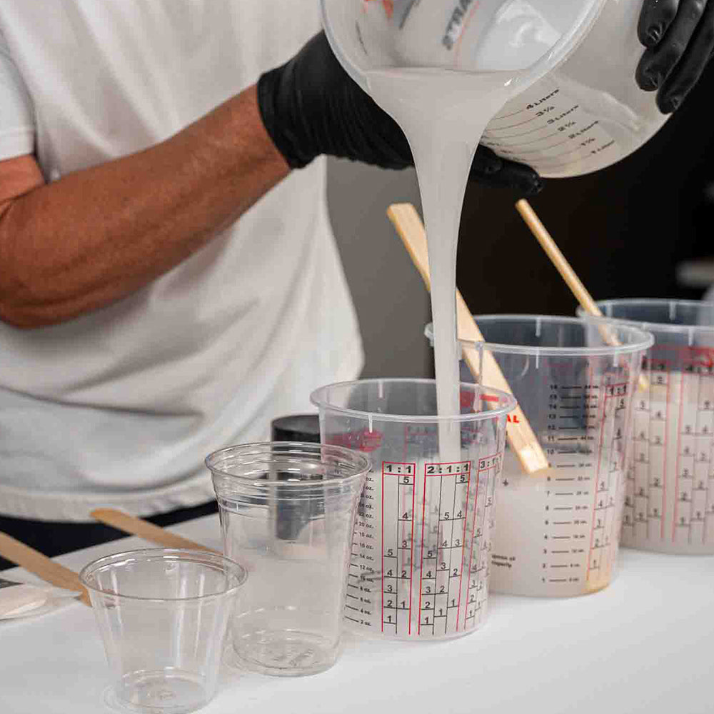 Person pouring APEX countertop white coating from a measuring cup into plastic containers with measurement markings.