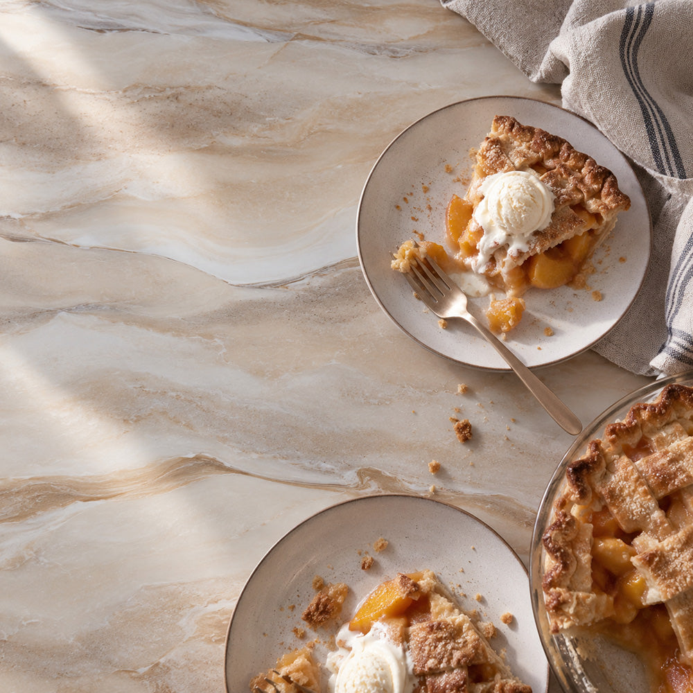 A styled overhead scene of peach pie served on white plates with a scoop of vanilla ice cream and a fork, alongside a glass pie dish on a light marble-like surface with a folded striped napkin.