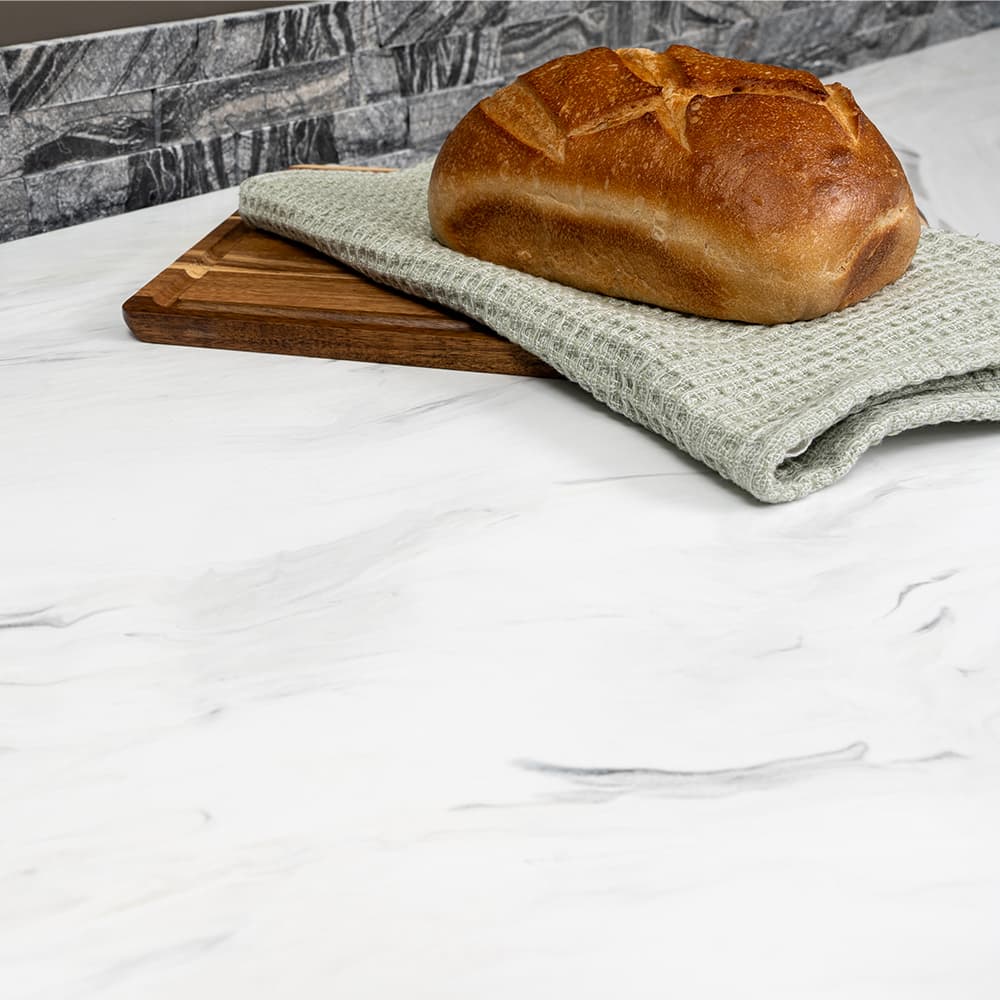 Loaf of bread on a wooden cutting board with a green cloth on a marble countertop.