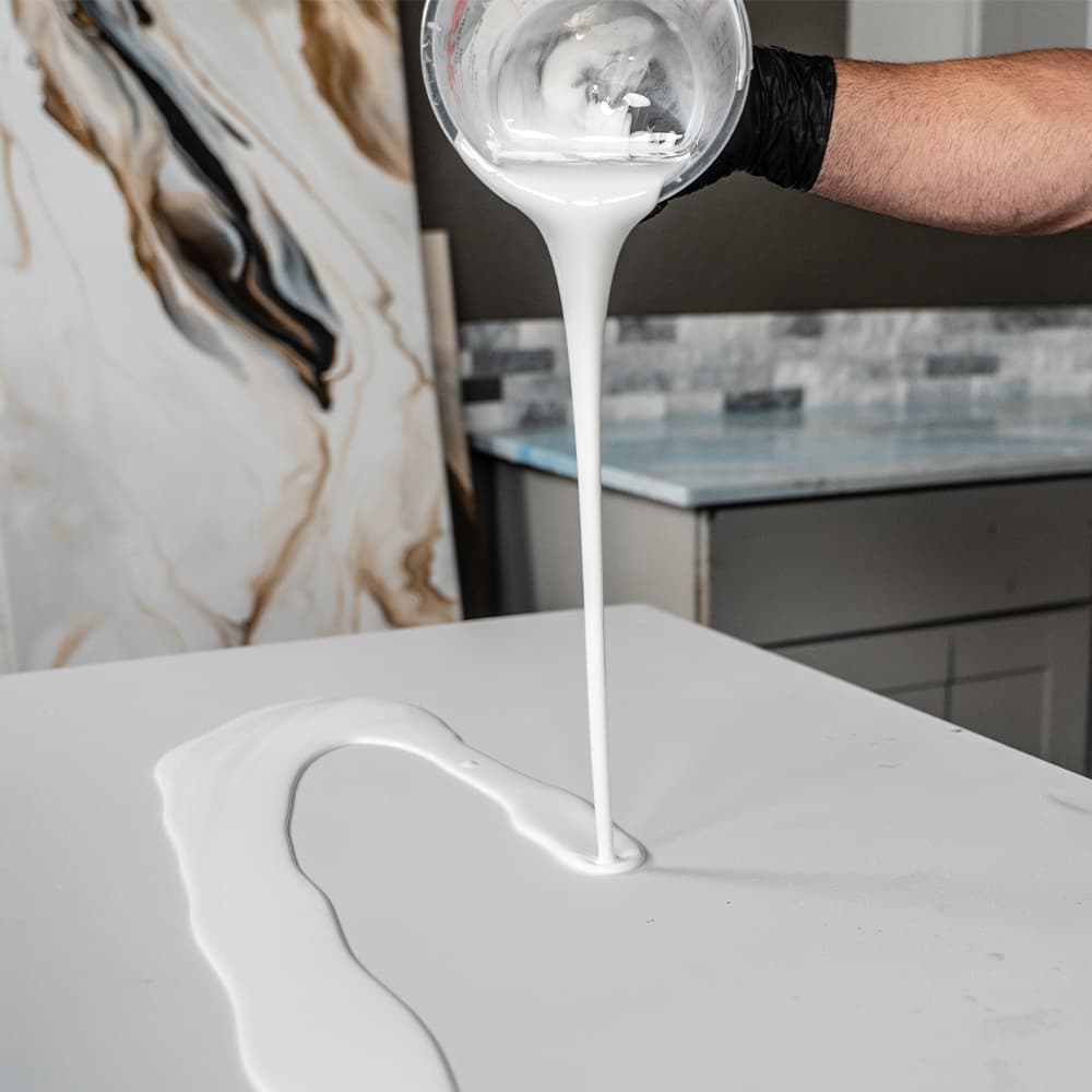 APEX white countertop dye being poured onto a white surface with a marble-patterned wall in the background.