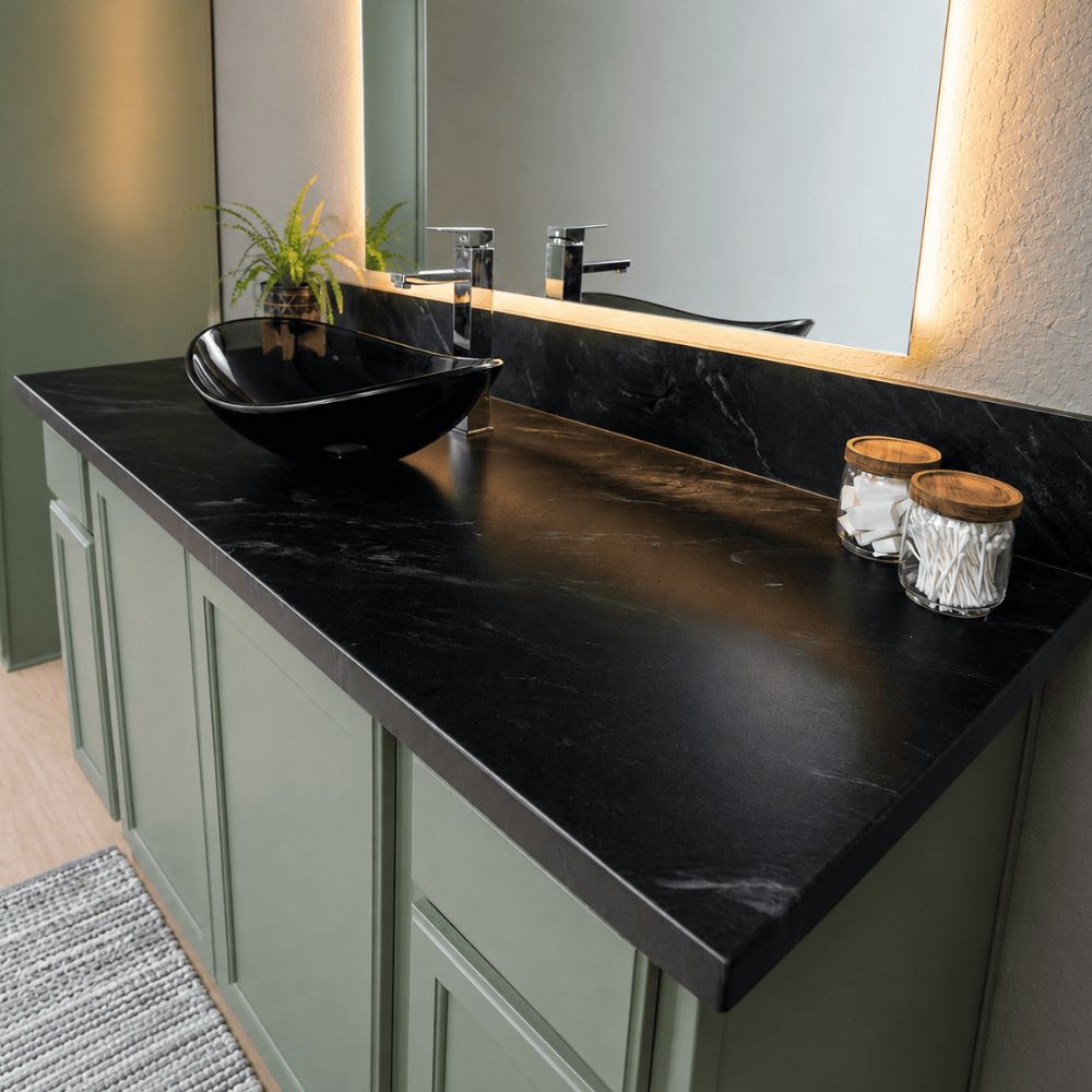 A modern bathroom vanity setup with a black stone epoxy countertop, black vessel sink, sage green cabinet doors, a large mirror with warm lighting, and small decorative jars on the counter.