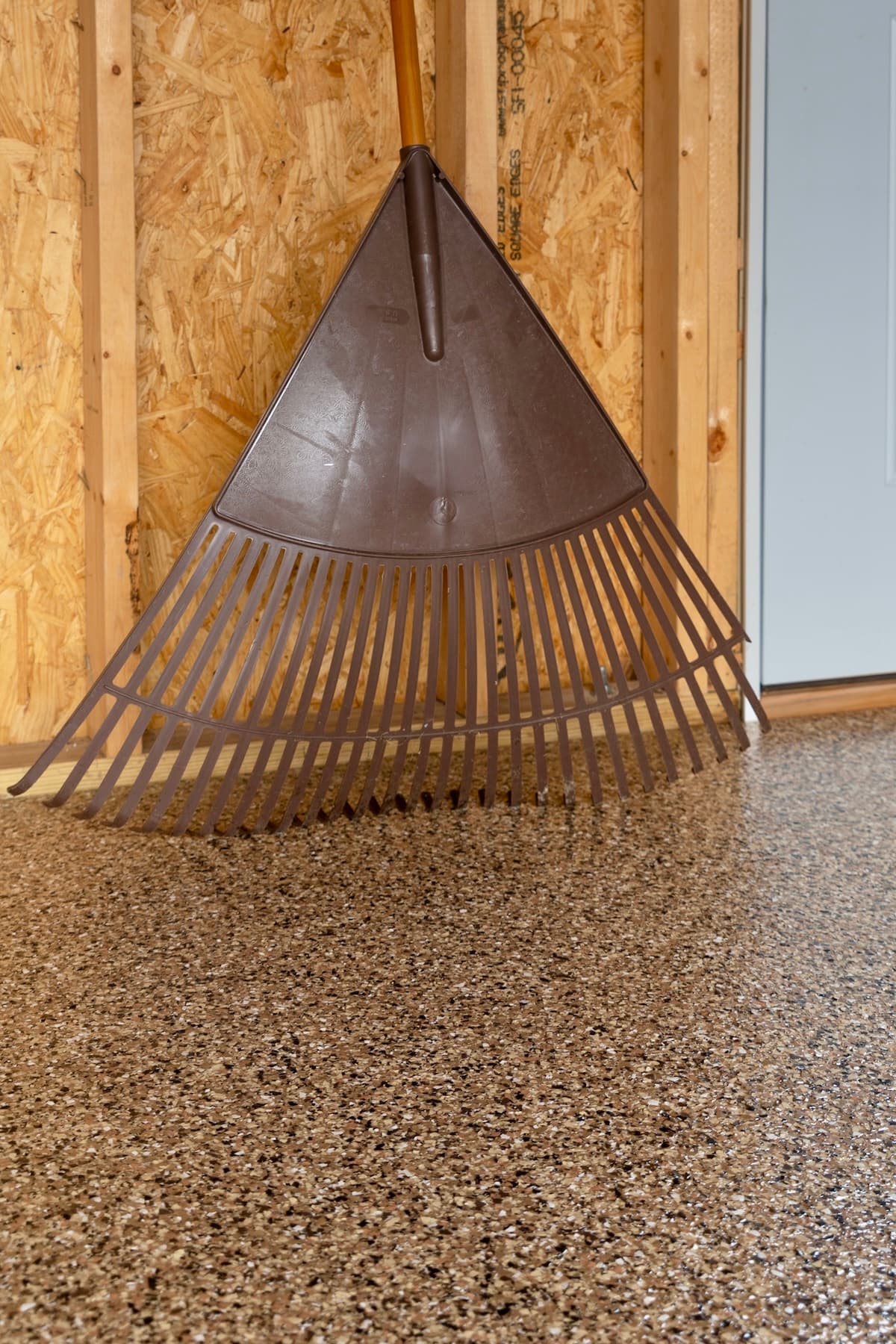 Rustic metal leaf rake leaning against a wooden wall in garage with brown epoxy floor flake.
