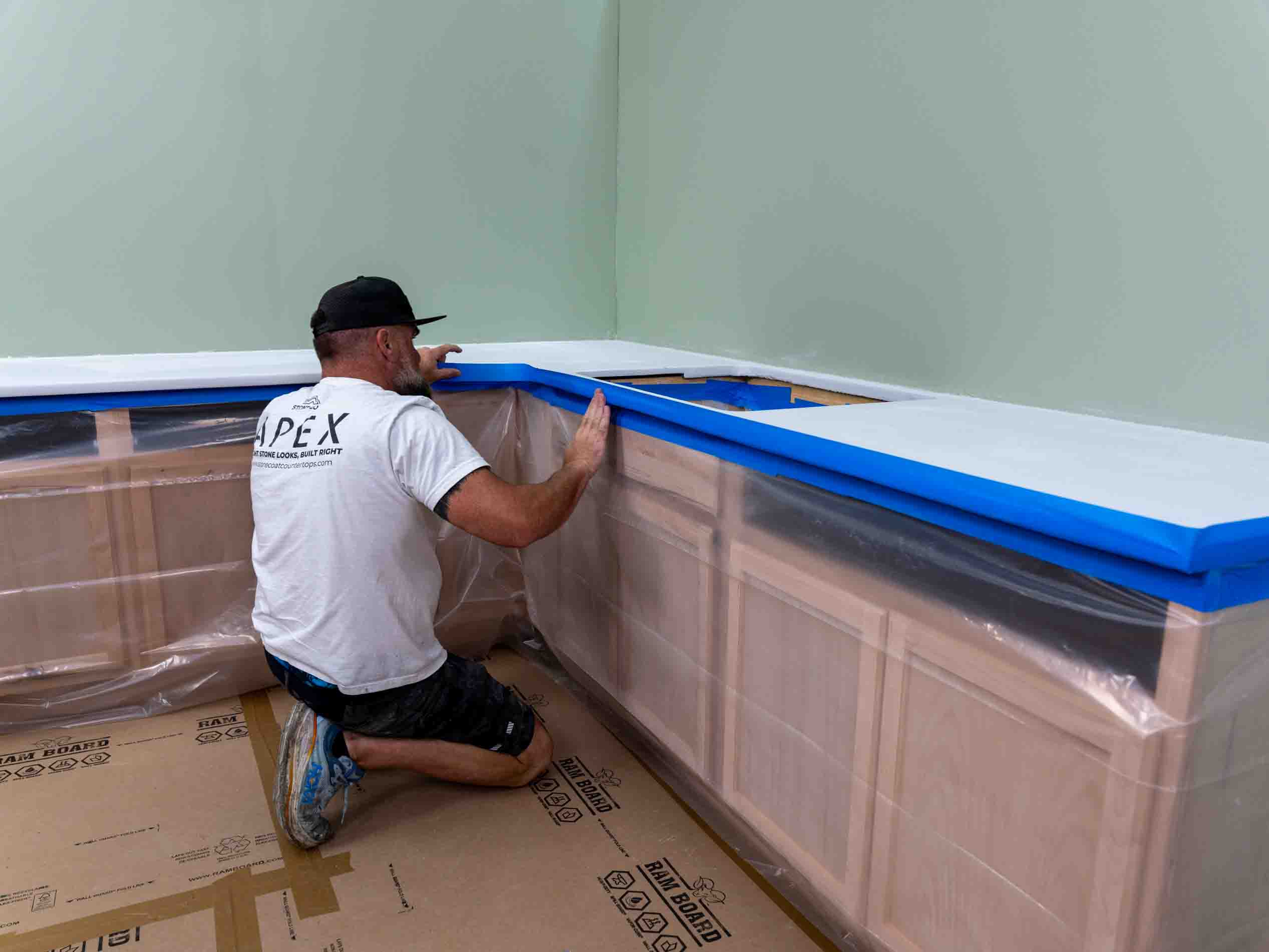 Applying protective plastic wrap to a light wood kitchen cabinet unit with a white countertop and creating blue tape dam along the edge.