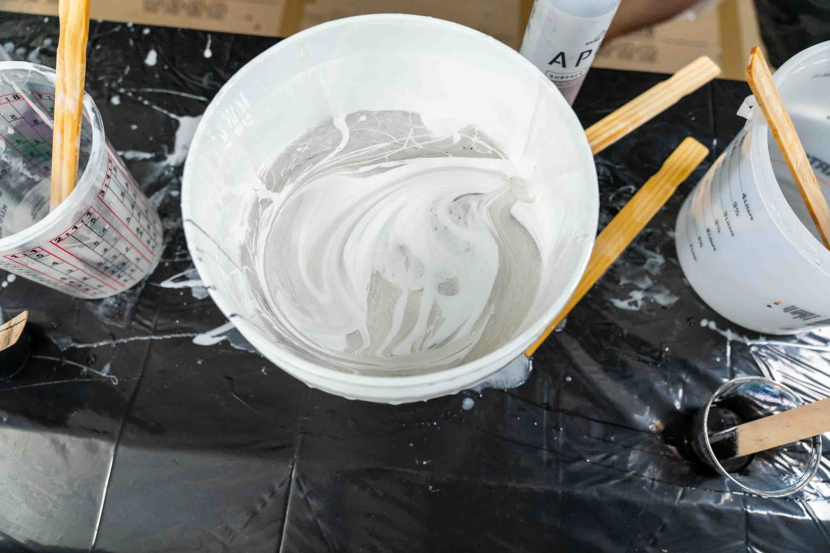A white plastic bucket filled with light gray to white powder or pigment slurry, surrounded by mixing cups, wooden sticks, and a black plastic sheet on a work surface.