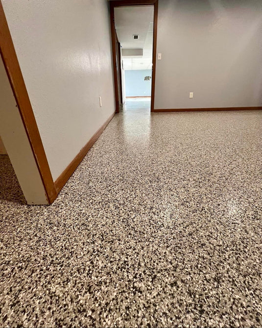 A wide view of a polished speckled terrazzo epoxy flake floor in a basement with off-white walls and wood trim, looking down a hallway toward an open doorway.
