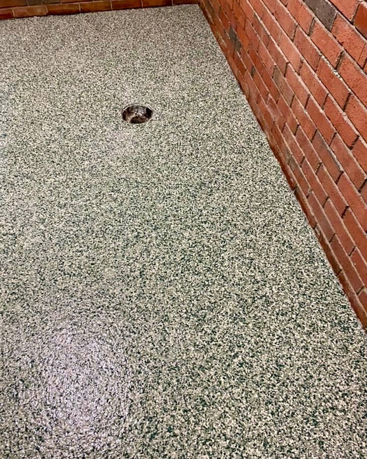 A close-up view of a speckled concrete epoxy floor coating in a garage or utility room corner, with a red brick wall on the right and a drain visible in the center.