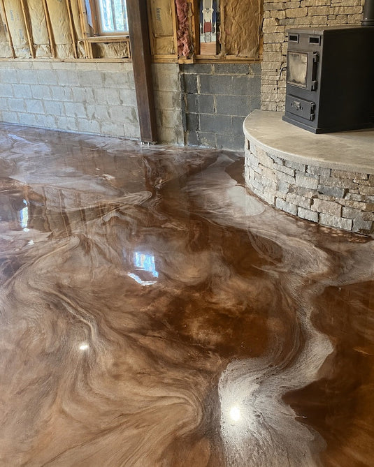 A glossy, swirling brown and beige metallic epoxy resin basement floor finish installed in an unfinished room with exposed wood framing, a stone wall, and a black wood-burning stove on a stone platform.