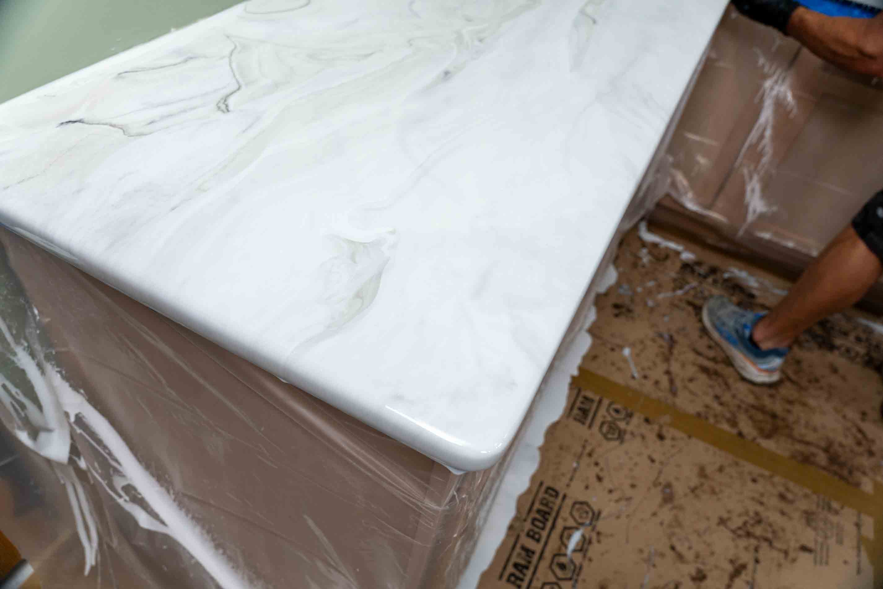 A white exotic marble-patterned countertop with a thick rounded edge, mounted on a light brown cabinet base and wrapped in protective plastic.